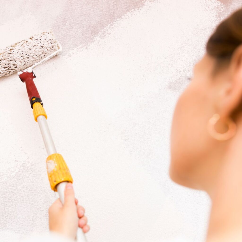 A woman from behind uses a roller to apply a paint swatch to a light gray interior wall.