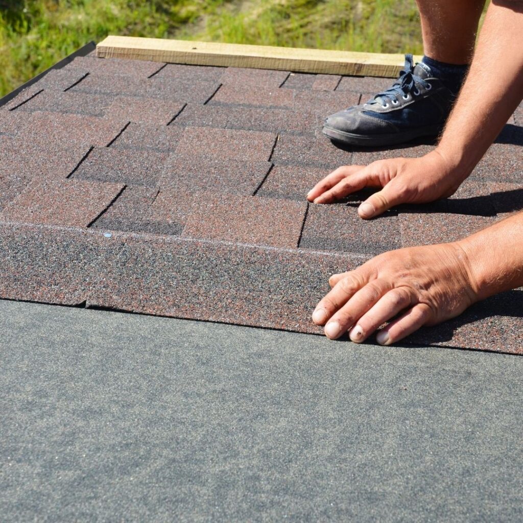 Hands laying reddish-brown asphalt shingles on a roof